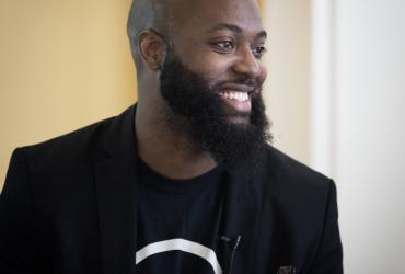 Nathan Holder is smiling and looking to the right. He has a beard and is wearing a black suit jacket over a black T-shirt with white text printed on it.