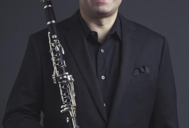 Peter Stoll is wearing a black suit, with a black background behind him. He is smiling and looking ahead, holding a clarinet in his hand.