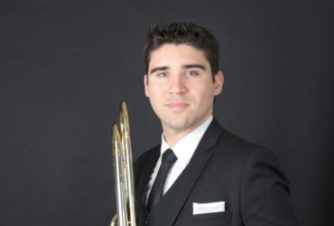 Trombonist Fabrice Millischer, against a black background, dressed in a black suit, wearing a tie, holding a trombone.
