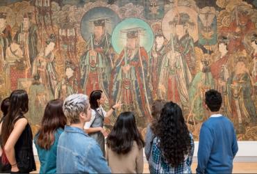 Tour Guide speaks to a group of 8 people standing in front of a large Chinese mural.